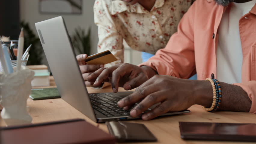 Close-up of hands of unrecognizable black couple purchasing vacation tickets online with credit card, man typing on laptop computer while woman reading aloud number