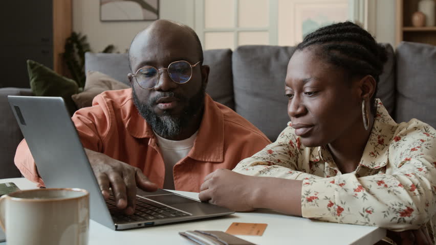 Medium close-up of African American husband and wife searching for travel options on laptop, choosing convenient flights for low price on booking aggregator website, wallet and credit cards on table