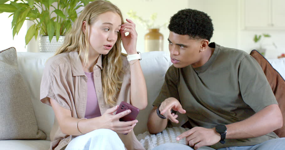 Couple on sofa, woman holding purple phone reacting to alert, man pointing, seeking explanation. Livingroom, couch, conversation, daylight, decor, plant, smartwatch