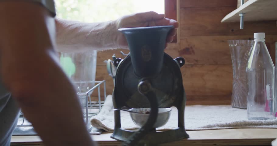 Man reaching to engage crank, turning grinder in kitchen, catching grounds while blue HUD growing. Castiron, manual, mill, countertop, wooden, rustic, vintage