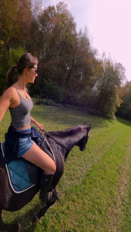 VERTICAL, SELFIE, LENS FLARE: Young woman in sunglasses rides a dark brown horse across a sunny grassy meadow beside fresh green forest. Horseback riding in beautiful spring natural environment.
