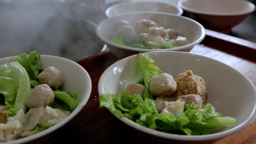 Steaming Asian meatballs and dumplings with green vegetables in a white bowl on a wooden table, close-up of traditional comfort food.
