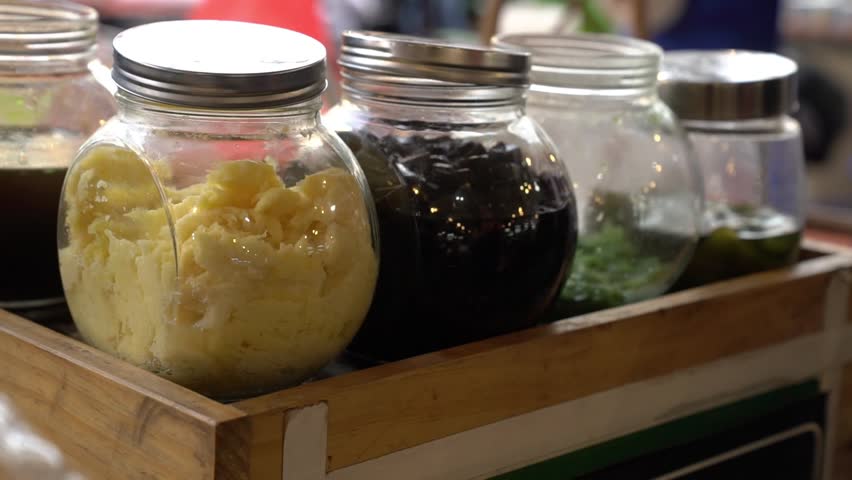 Glass jars filled with street-side mixed ice toppings on a wooden cart, colorful dessert ingredients at a warm market, close-up.