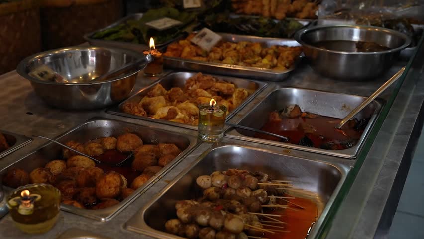 Warm Indonesian street food buffet with fried sides, eggs, skewers, and spicy dishes in stainless trays at a traditional food stall.