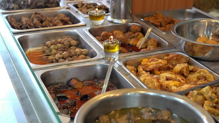 Authentic Indonesian food buffet with assorted traditional dishes in stainless trays, warm street food display, rich local cuisine.
