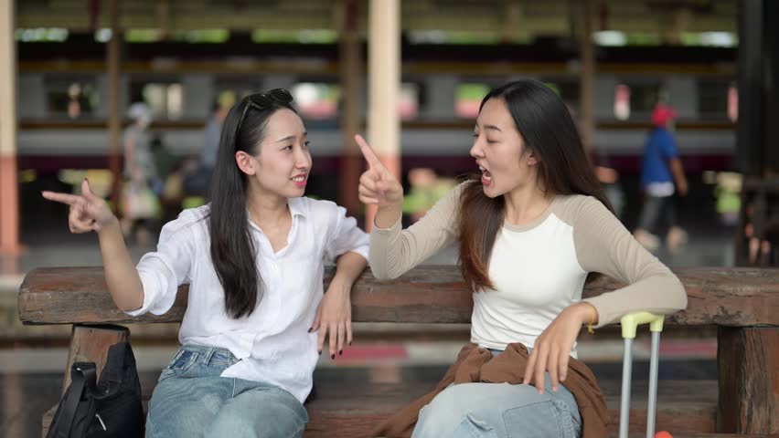 Two women sitting on a bench, one pointing at something. Scene is lighthearted and playful