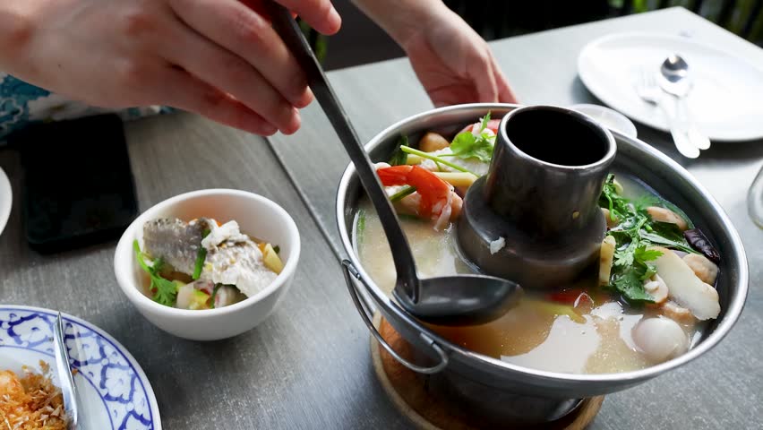 A person ladles hot seafood soup from a traditional metal pot into a small bowl