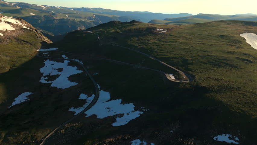 Beartooth Highway in Montana curves dramatically through high alpine terrain with patches of melting snow dotting green meadows. Golden hour light illuminates the scenic mountain pass landscape.