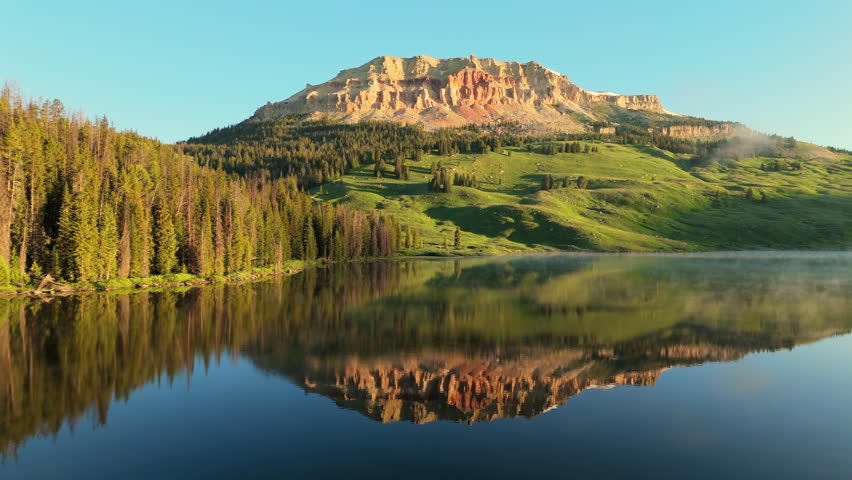 Stunning mountain peak with red rock cliffs reflects perfectly in glass-like alpine lake water. Morning mist rises over evergreen forest as golden sunrise light illuminates the rugged landscape.