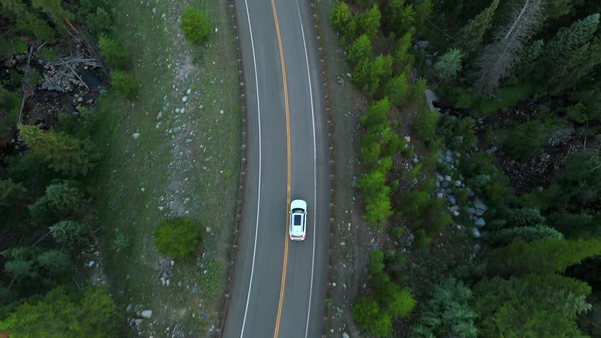 Cinematic aerial white car traveling along winding mountain highway through lush green Montana forest. Pine trees and rocky terrain surround the scenic two-lane asphalt road.
