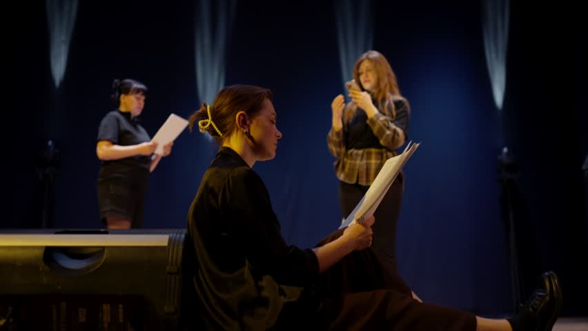 Aspiring actors rehearsing dramatic script on dimly lit theater stage, seated and standing performers practicing lines with papers under focused stage lighting