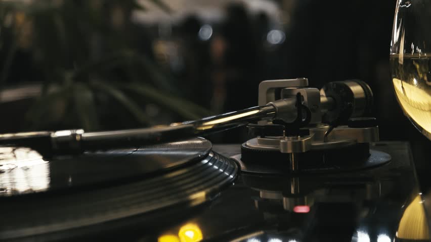 A vinyl record spins on a turntable with a stylus resting on the grooves, captured with a shallow depth of field and warm bokeh lights in the background.