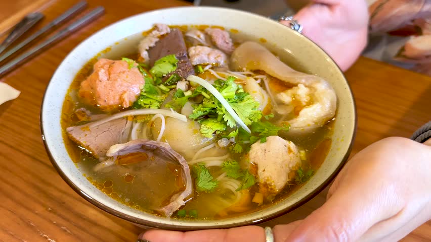 Hands rotate a bowl of spicy Vietnamese beef noodle soup on a wooden restaurant table