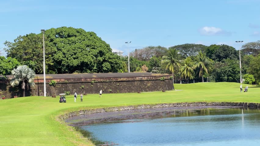 Golf course, lake, ancient wall, sunny day, golfers, peaceful nature.