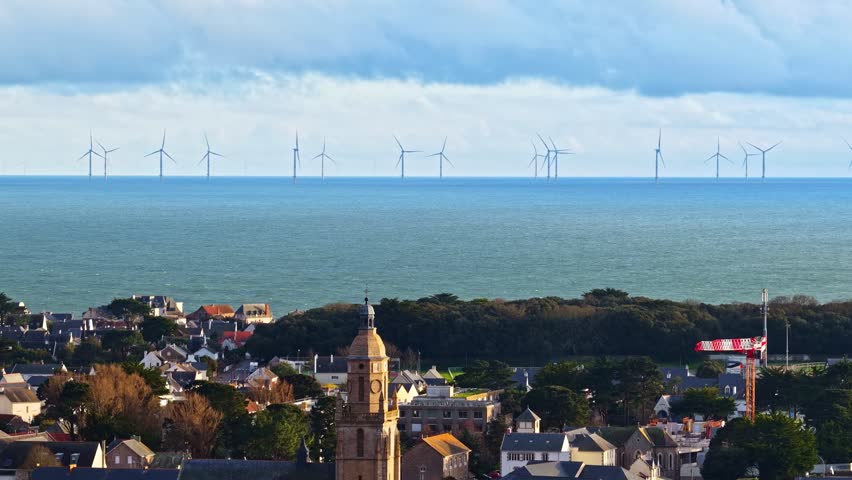 A coastal village with a prominent church tower overlooks the sea, where wind turbines line the horizon. Ideal for projects on renewable energy or coastal communities.