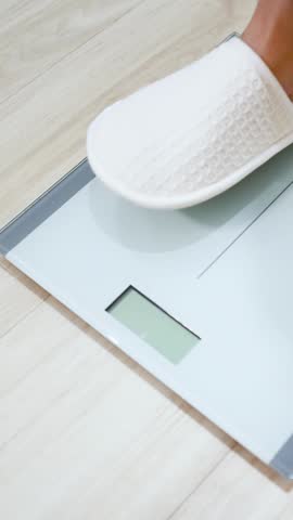A high-angle shot of feet in slippers stepping onto a digital scale on wooden flooring