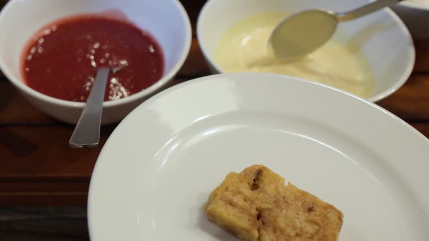 A person pours creamy vanilla sauce from a spoon onto bread pudding in a restaurant