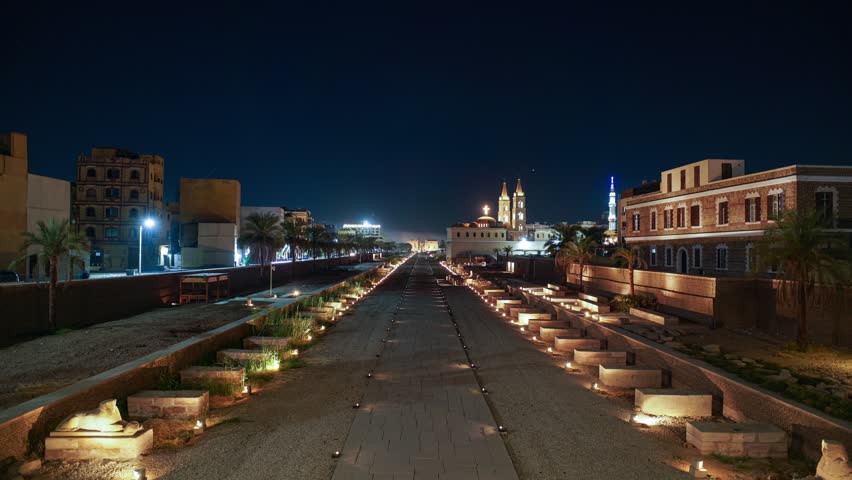 Night time lapse of illuminated Avenue of Sphinxes leading toward Luxor temple in Egypt, flanked by palms, historic architecture ruins and church under a clear starry sky.
