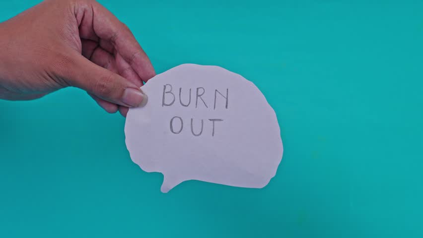 Hand holds pink paper brain cutout with handwritten words "BURN OUT" as it is set on fire by match in slow motion