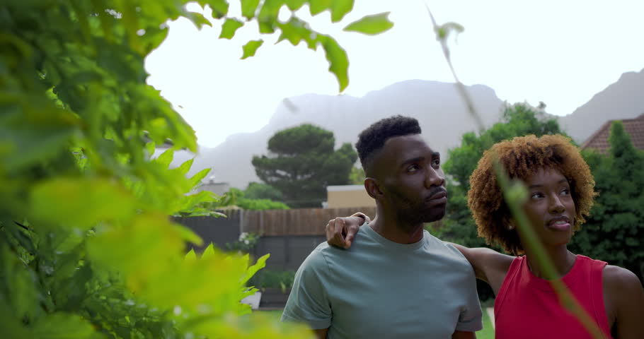 Couple standing in backyard by leafy shrub, man spotting then showing view, pointing, woman smiling. Pair, outdoor, pool, lawn, fence, mountains, candid