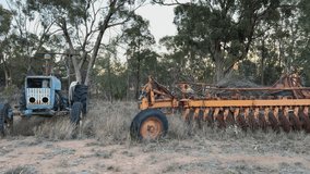 A vintage blue tractor sits idle beside rusty orange disc harrow farming equipment in the Australian outback, surrounded by eucalyptus trees and dry grass in the fading afternoon light. - Powered by Shutterstock - Get 15% off with code: PIKWIZARD15