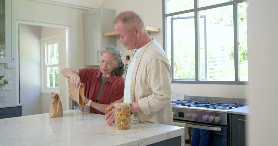 Senior woman pouring legumes into jar, senior man placing lid, organizing pantry at island. Couple, kitchen, modern, bright, storage, organization, shelves
