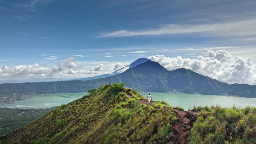 Athletic man running mountain ridge overlooking Lake Batur and volcanic Mount Agung peak under blue sky. Active sport, challenge and recreation. Travel and explore beautiful nature of Bali, Indonesia