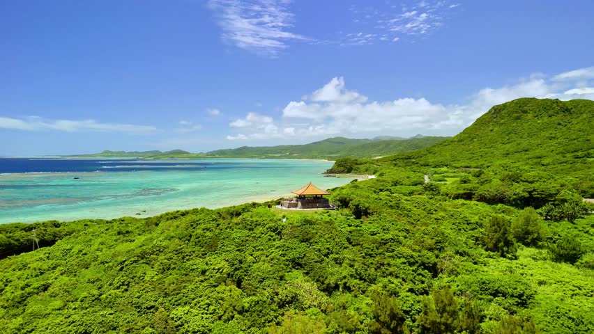 Cinematic drone shot of Tamatorizaki Observatory overlooking the turquoise coral reefs and lush tropical mountains of Ishigaki Island, Okinawa, Japan