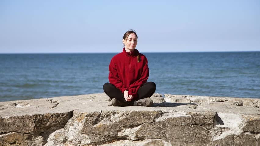 Peaceful woman sitting cross-legged on large rocks by calm blue sea wearing vibrant red sweater during bright daylight. Relaxed contemplation with minimal background and natural light