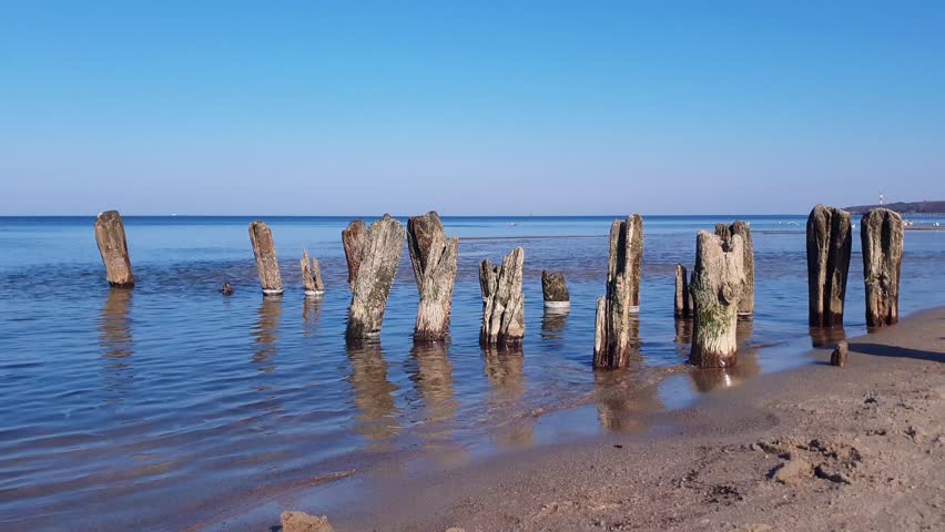 Tranquil seaside landscape of Baltic sea coast with calm water waves rolling on sand beach and old damaged wooden breakwater under clear blue sky at spring day. No people seascape natural background.