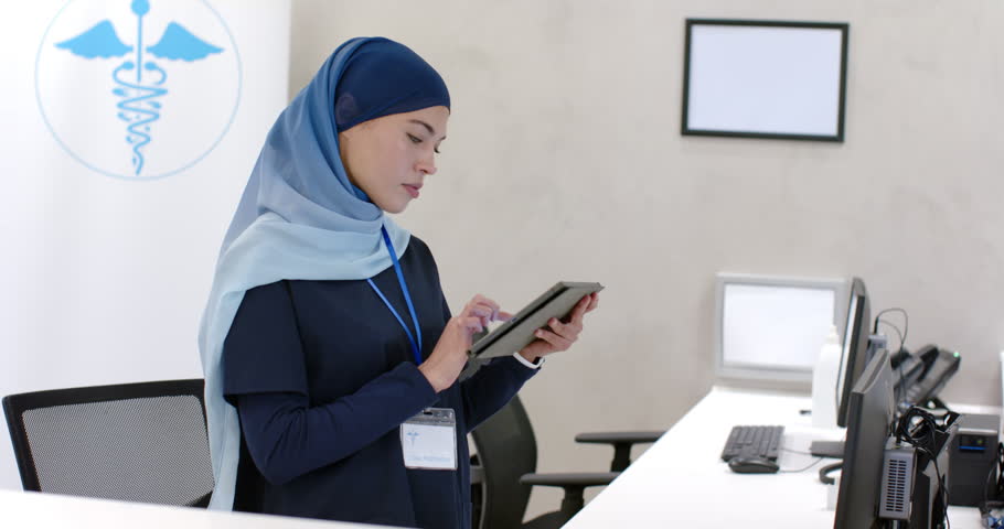Woman in navy scrubs hijab tapping and scrolling tablet after check-in prompt at clinic desk. Healthcare, receptionist, touchscreen, technology, professional, office, efficiency