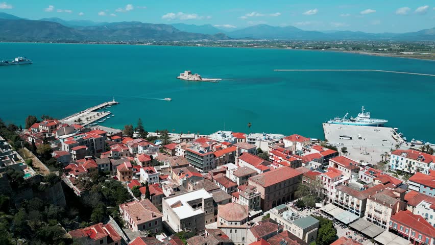 High-angle aerial view of Nafplio, a historic coastal city in Greece, featuring traditional red-tiled rooftops, the deep blue waters of the Argolic Gulf, and the Bourtzi water fortress located on a small island in the harbor. Smooth cinematic drone rotation over the old town and waterfront on a bright sunny day. Scenic Mediterranean landscape with clear sky, calm sea, and classic European architecture. Ideal for travel, tourism, summer vacation, and destination content.
