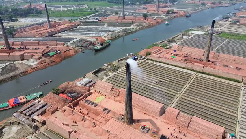 Aerial view of traditional brick kilns with tall chimneys emitting smoke and stacks of red bricks along a river Narayanganj, Dhaka Division, Bangladesh.