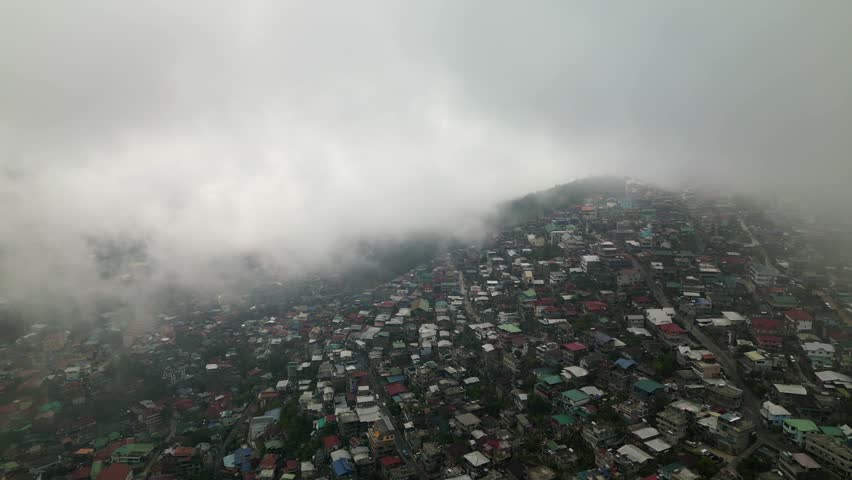 Aerial orbit circling a dense hillside community with colorful rooftops and fog drifting across Quirino Hill, Baguio City, Philippines