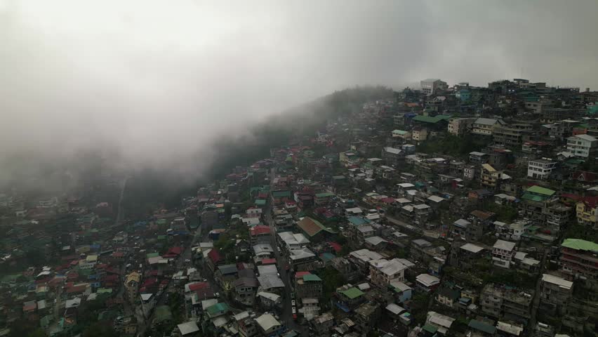 Aerial flyover of a densely populated hillside barangay with compact homes and fog drifting across Quirino Hill, Baguio City, Philippines