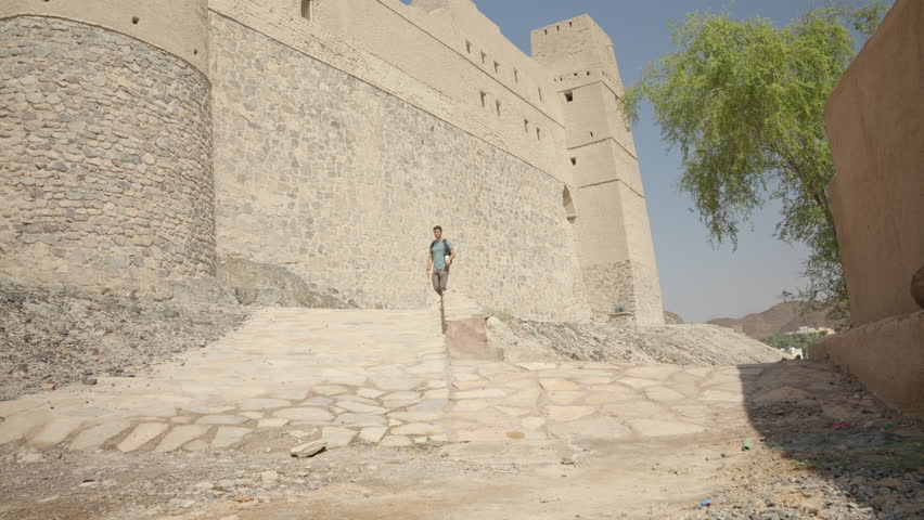 Horizontal static shot of a male tourist walking alongside the massive unbaked mud-brick walls of Bahla Fort. Showcasing the scale of the ancient defensive towers in the Omani desert.