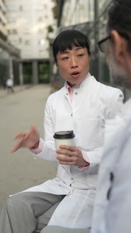 Two medical professionals, a man and an asian woman, are having a conversation during a coffee break outside the hospital building, with the woman actively gesturing while speaking to her colleague