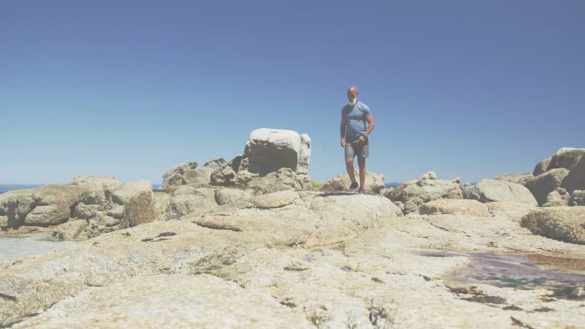 Vertical video: HUD appearing, man jogging down rocks toward lens in tshirt shorts shoes, padlock. Male, hiker, coastline, shoreline, boulders, tidepool, outdoor