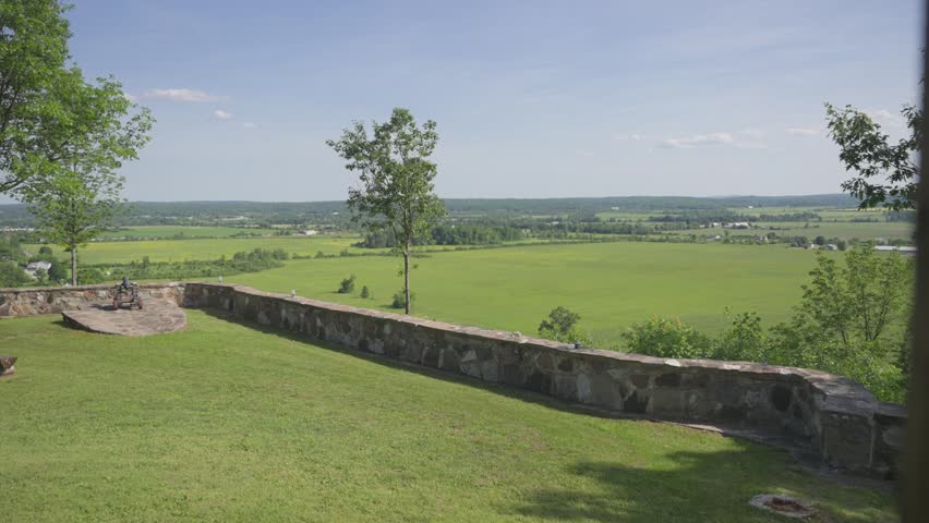 A wide-angle panoramic view from a stone-walled lookout featuring a historic cannon, overlooking vast green fields, rural farms, and distant hills under a clear blue sky.