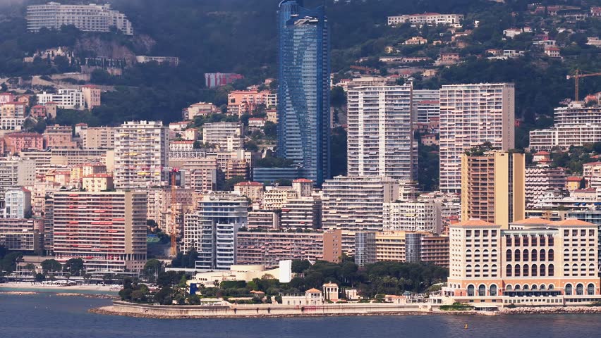 Aerial view shows Monaco skyline on the French Riviera as the camera pans, a blue glass tower, terraced apartments, pastel villas, seawall, beach, boats, and a work barge.
