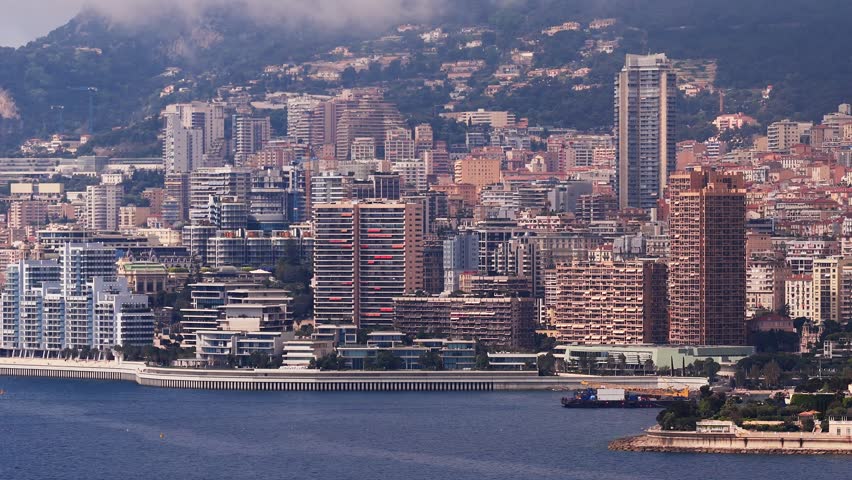 Aerial view of Monaco on the French Riviera shows Monte Carlo Casino, high rise towers, seawall, promenade, and a moored workboat, in warm hazy daylight.