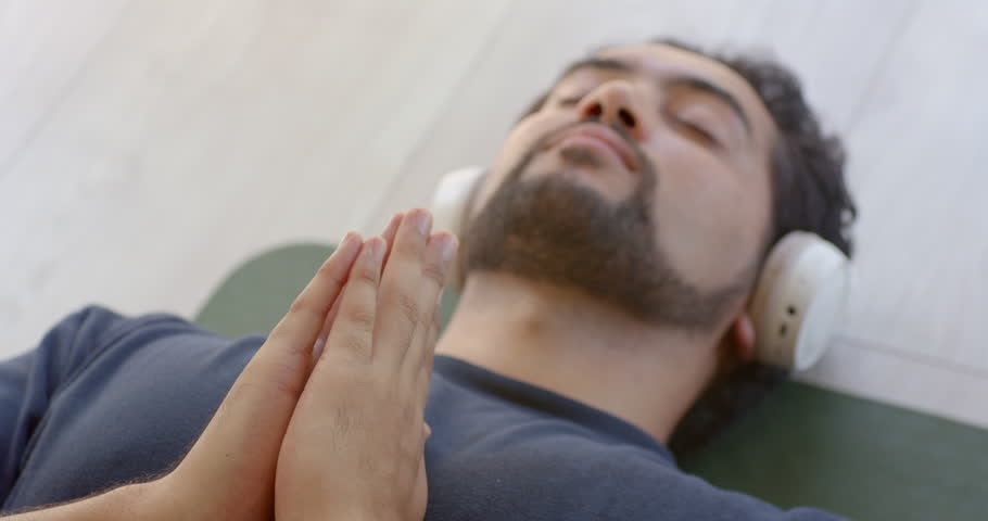 African man lying on green mat wearing white headphones praying hands breathing causing faint rise. Meditation, mindfulness, relaxation, yoga, serene, closeup, earphones