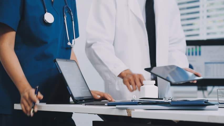 A vibrant red anatomical heart model and a stethoscope on a desk with two doctors having a medical consultation in the background.