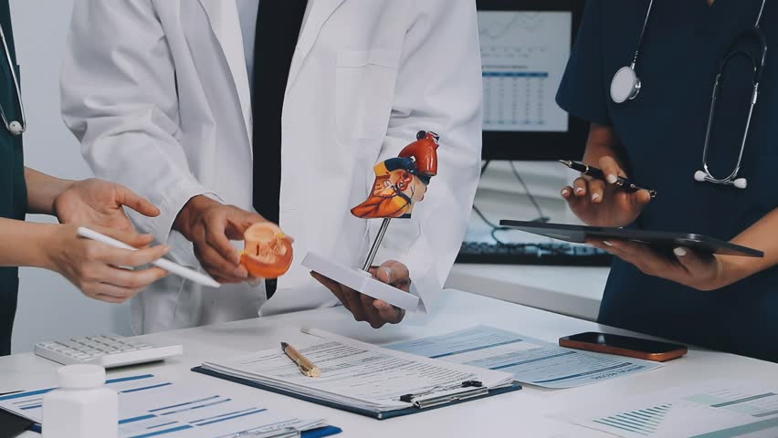 A vibrant red anatomical heart model and a stethoscope on a desk with two doctors having a medical consultation in the background.