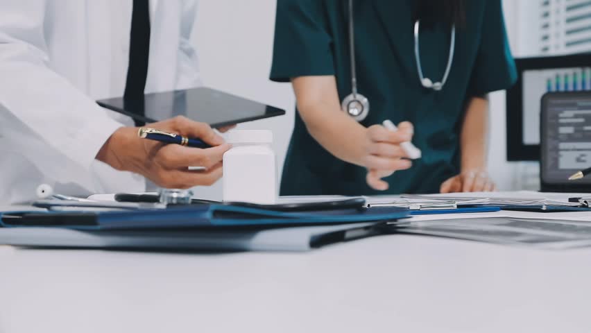 A vibrant red anatomical heart model and a stethoscope on a desk with two doctors having a medical consultation in the background.