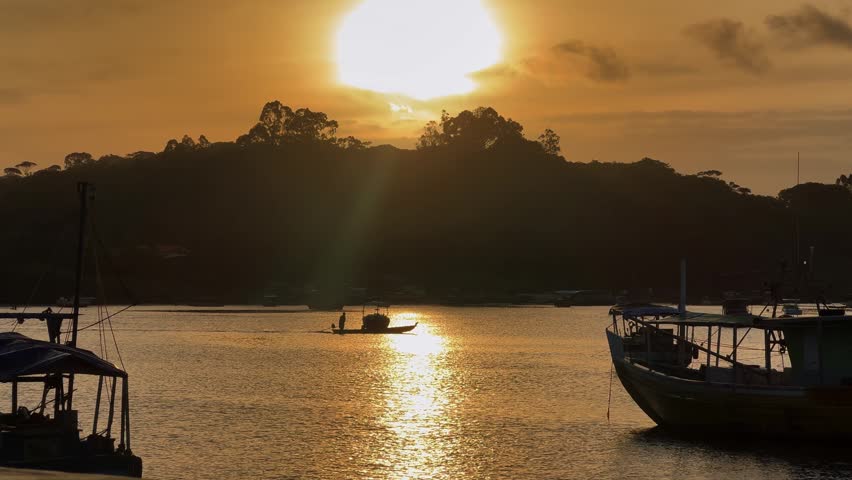 Golden sunset over the river in Itacaré, Bahia, Brazil, with a small boat and silhouettes of fishermen reflected on the calm water.