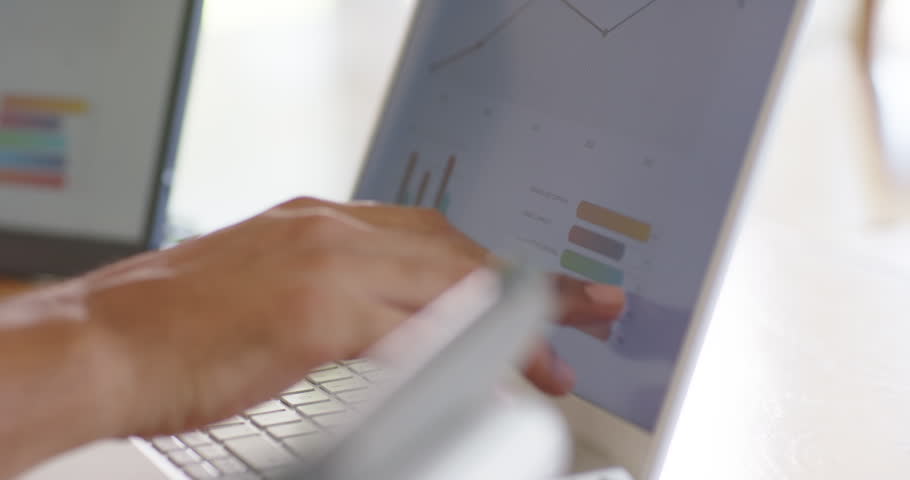 Right hand is typing and tapping laptop and monitor while responding to chart updates on desk. Keyboard, computer, multiscreen, dashboards, analytics, data, focus