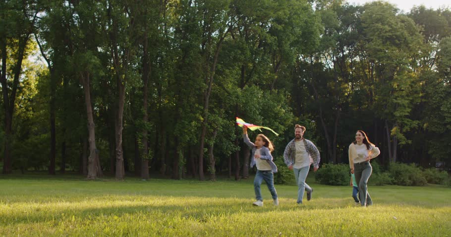 Portrait of happy family running on green grass in the park and flying kite together at sunset. Young parents with kids walking in sunny summer park outdoors. 4k video. Slow motion video.