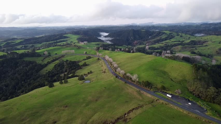 Drone aerial view of rural road passing through green farmland and rolling hills in New Zealand.