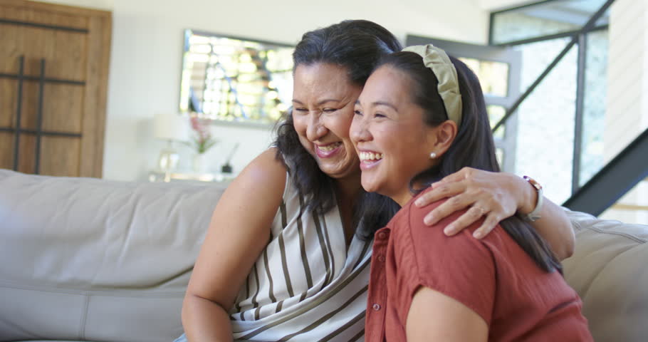 African American mom putting arm around Asian daughter making laugh and posing for camera on sofa. Family, mother, women, hug, smile, joy, affection